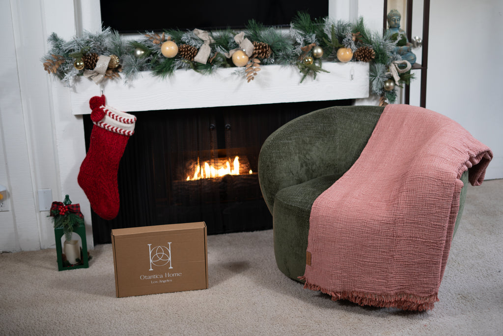 A blush pink muslin blanket draped over a green chair in front of a decorated Christmas fireplace, showcasing soft textured fabric and cozy holiday ambiance.