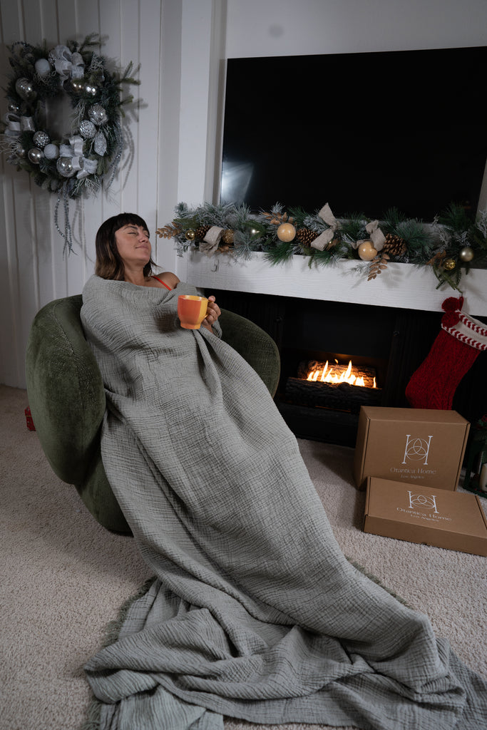A sage muslin blanket styled over a chair with soft folds, displayed in a festive room with holiday decorations.