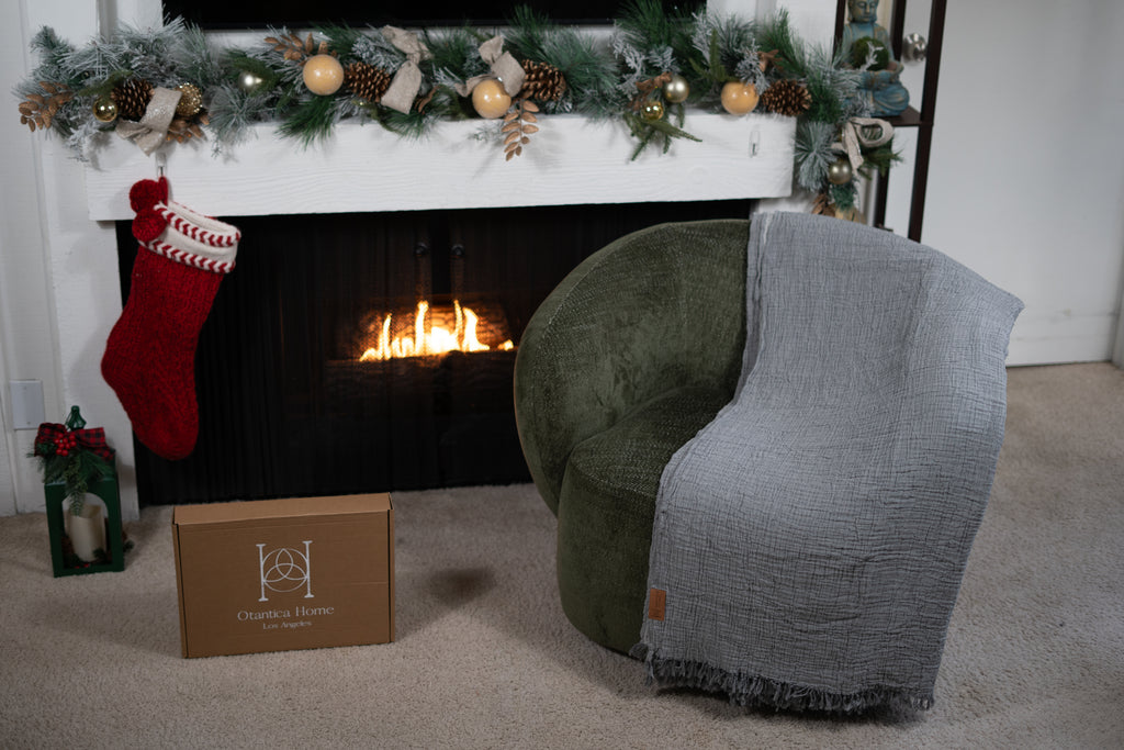 A grey muslin blanket draped over a green chair next to a fireplace, showing textured double-gauze organic cotton fabric.
