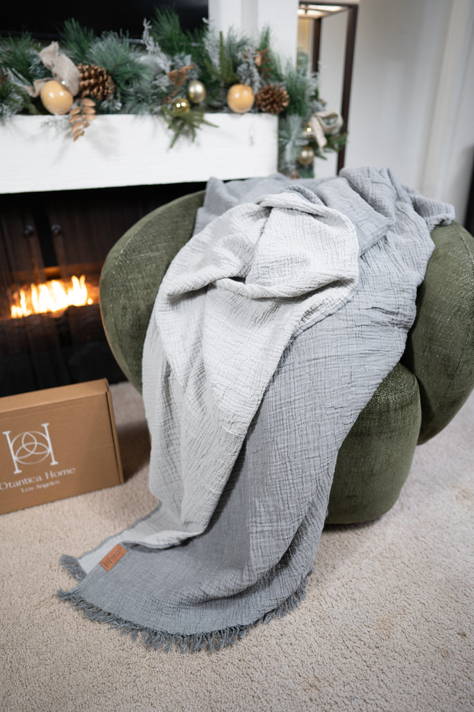 A sage muslin blanket draped over a green chair with Christmas garlands and a lit fireplace in the background.