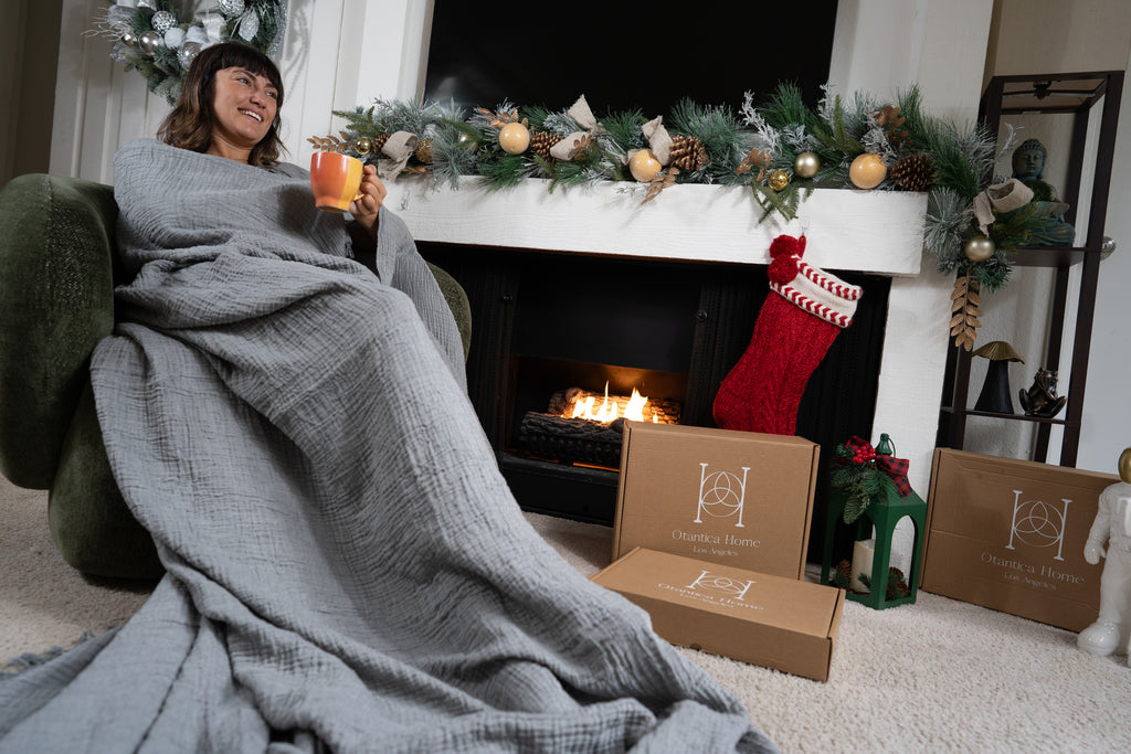 A woman lounged comfortably in a grey muslin blanket, smiling while holding a warm mug near the fireplace.