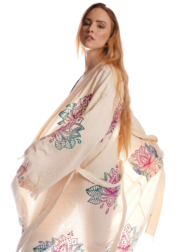 A woman wearing the Organic Lotus Kimono by Otantica Home, showcasing multicolor hand-embroidered lotus floral patterns on a beige organic cotton kimono, standing against a white background.