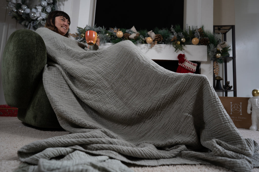 A woman wrapped in a sage muslin blanket enjoys a warm drink while relaxing near the fireplace in a cozy holiday environment.