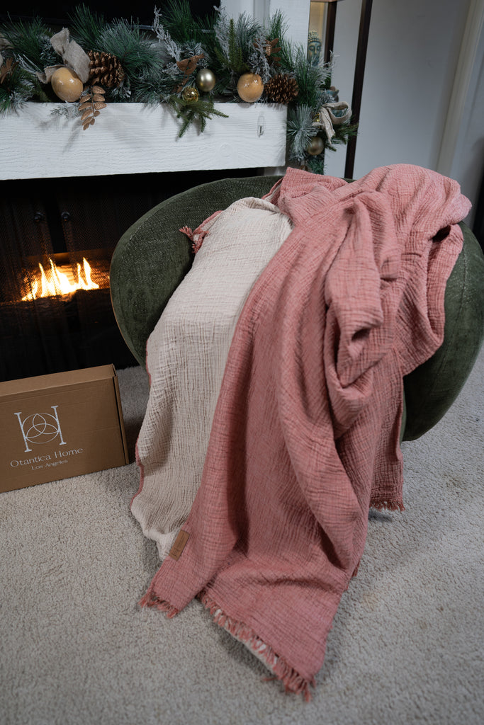Blush and sand muslin blankets layered over a green chair near a fireplace, highlighting double-gauze texture and organic cotton weave.