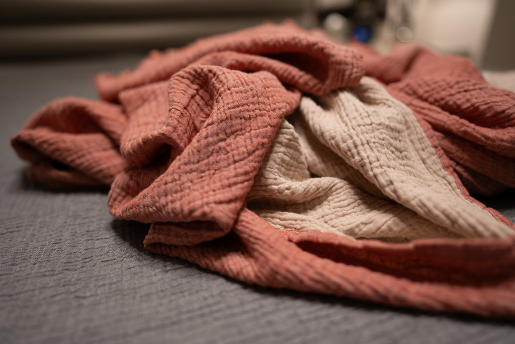 Close-up of blush and sand muslin blanket showing double-gauze crinkled texture and reversible soft cotton fabric.