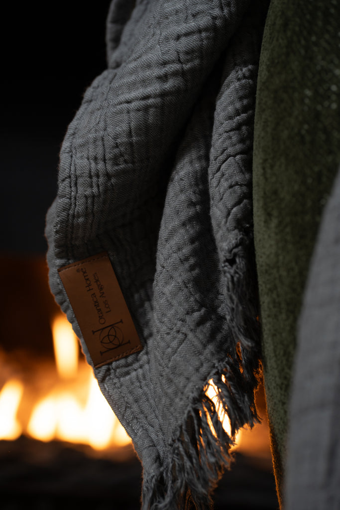 Close-up of the grey muslin blanket’s fringed edge and textured weave, illuminated softly by the fireplace glow.