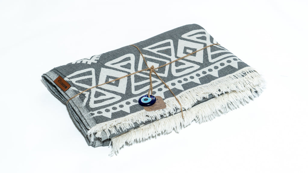 Folded grey boho blanket with fringe and evil eye charm – Otantica Home organic cotton.