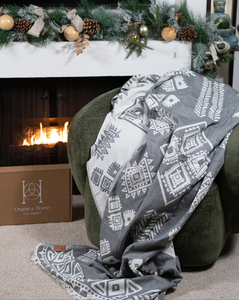 Grey organic cotton boho blanket draped over chair in front of fireplace – Otantica Home.