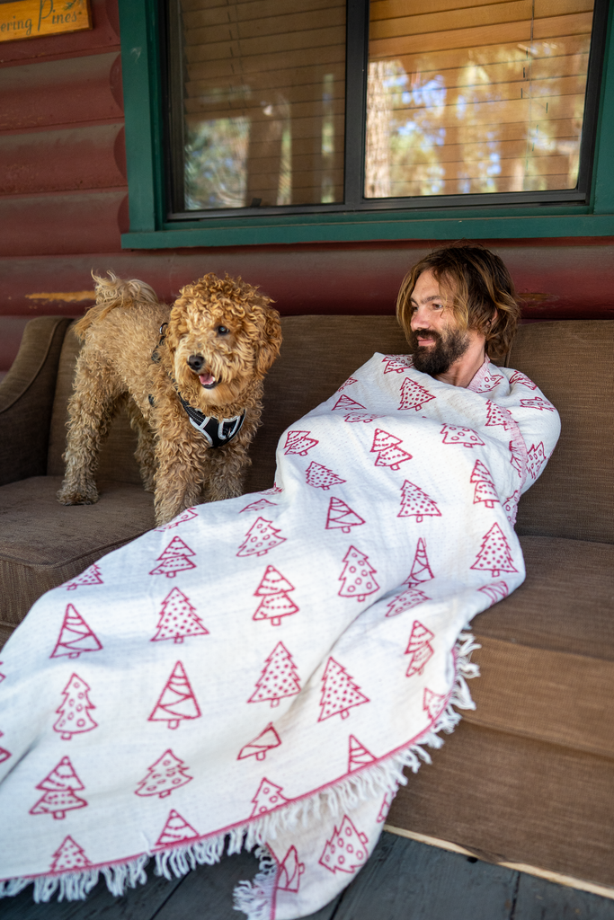 Man and dog relaxing on a porch with a white-and-red pine tree Christmas blanket, reversible organic cotton holiday throw.
