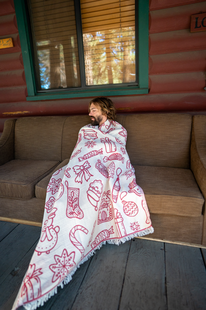 Man relaxing on a porch sofa wrapped in a red and white Christmas blanket, showing holiday-themed patterns.