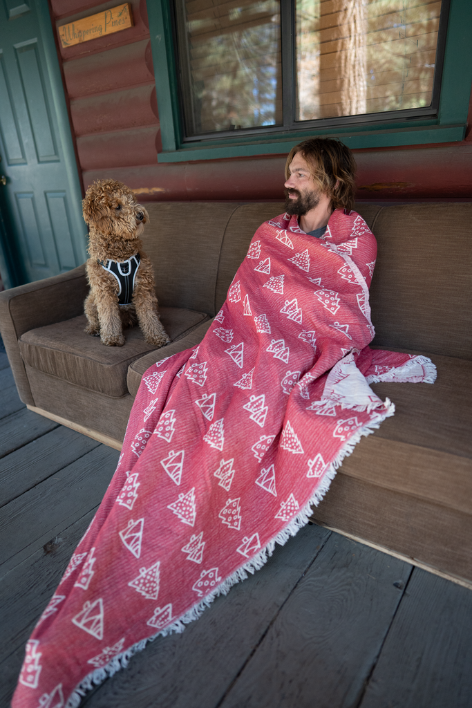 Man sitting on a cabin porch couch wrapped in a red pine tree patterned Christmas blanket with a dog beside him.