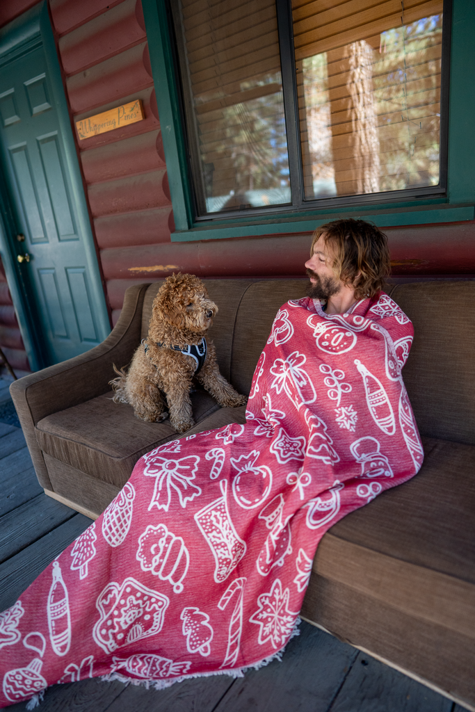 Man sitting on a cabin porch wrapped in a red and white Christmas blanket, with a dog beside him.