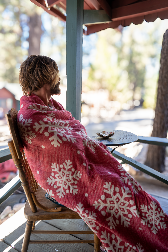 Man wrapped in Otantica Home Xmas Snowflake Blanket — red organic cotton Christmas throw with white snowflakes, cozy winter porch setting.