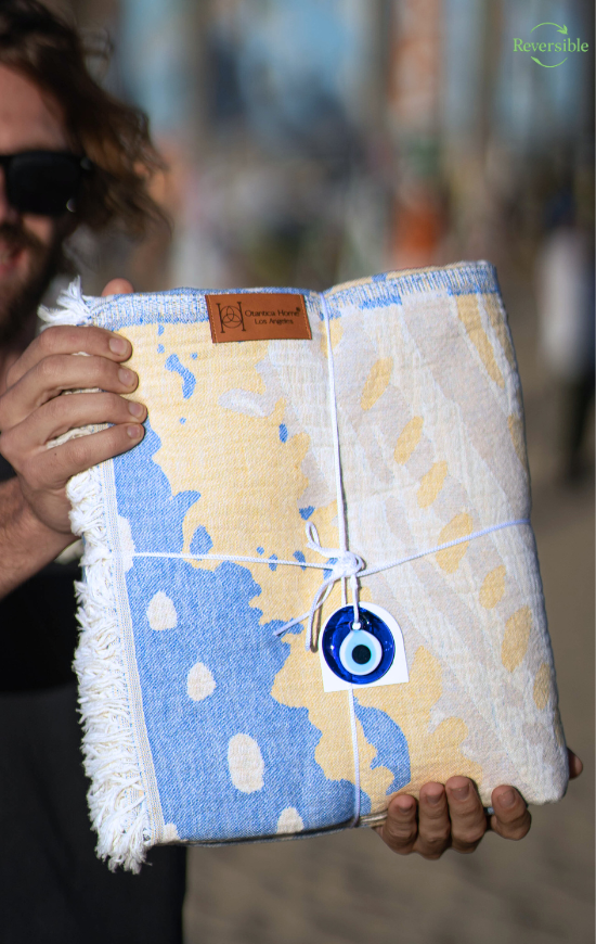 Person holding a folded blue and beige reversible Havana blanket with an evil eye charm tag.