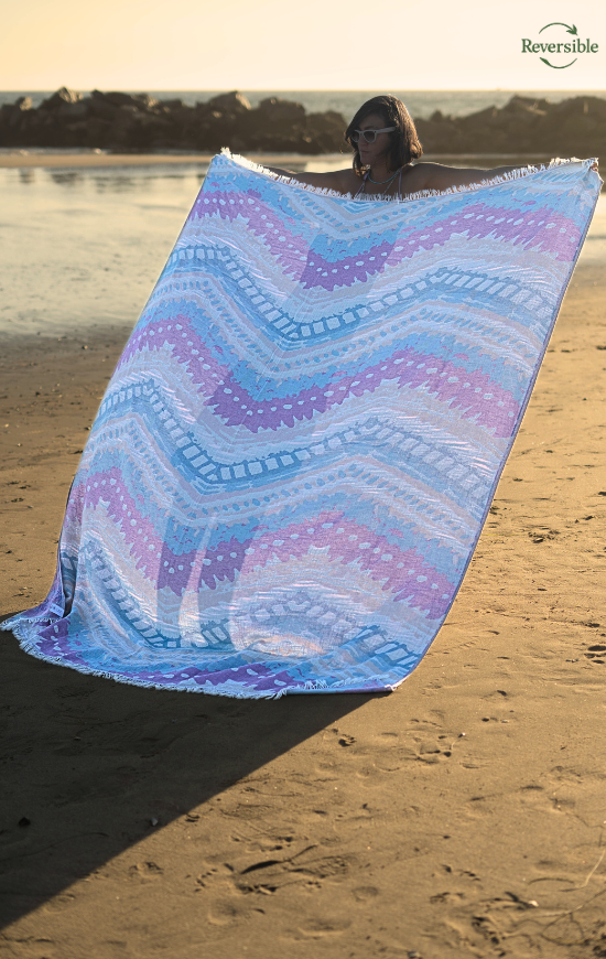 Person holding a large reversible Havana blanket in blue and purple pastel colors on a sunny beach.