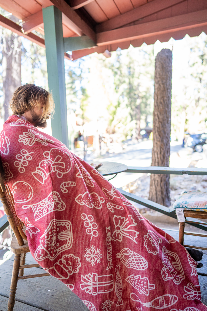 Person sitting on a wooden chair on a cabin porch, wrapped in a red and white Christmas blanket with festive patterns.