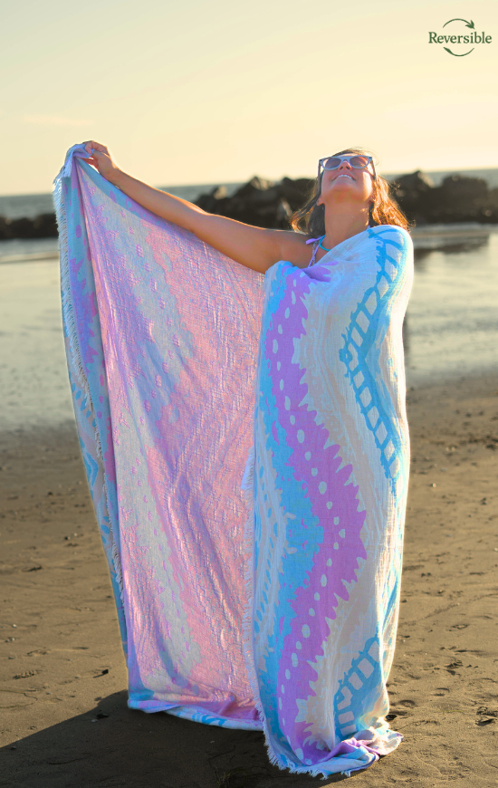 Person wrapped in a colorful reversible Havana blanket (blue, lavender, pink) on a beach at sunset.