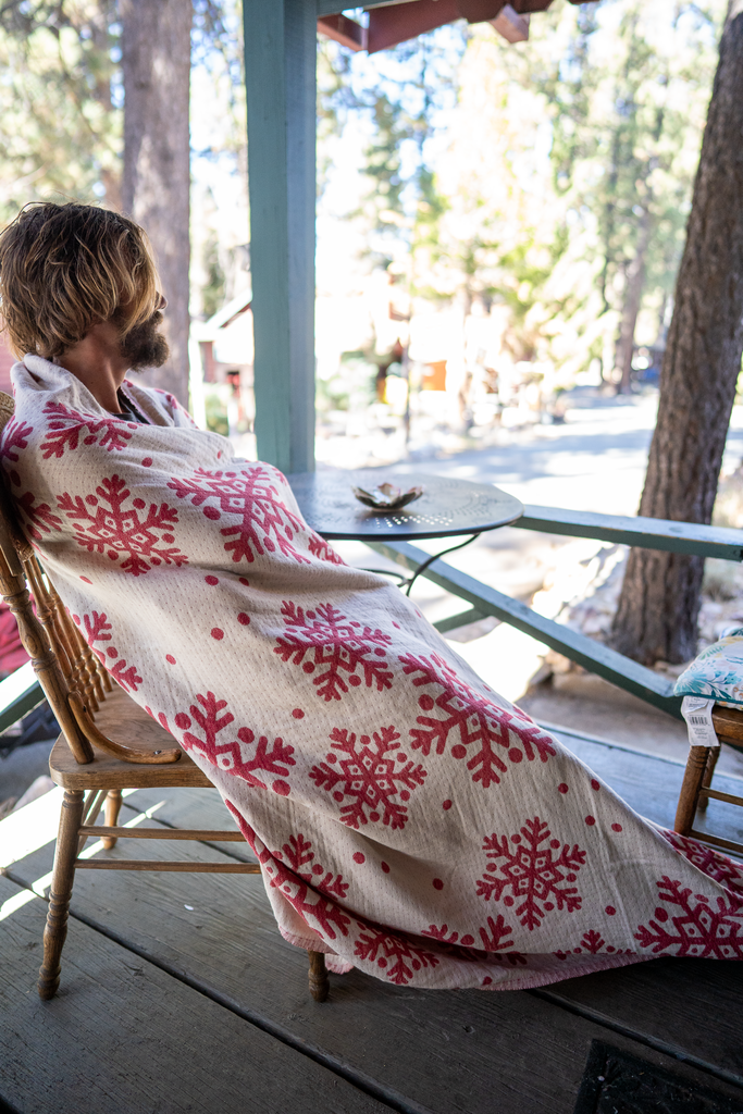 Reversible organic cotton Xmas Snowflake Blanket in red & white — person sitting on porch wrapped in holiday blanket, winter cabin atmosphere.