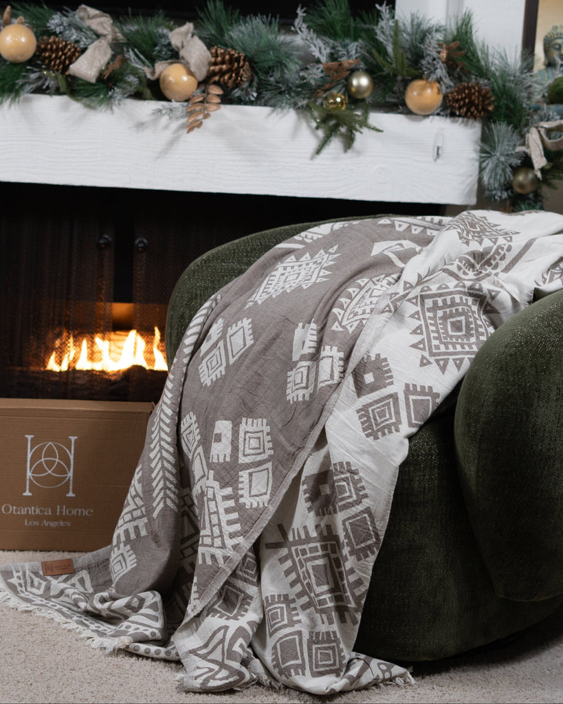 Sand boho organic cotton blanket draped over an armchair next to an Otantica Home gift box by the fireplace