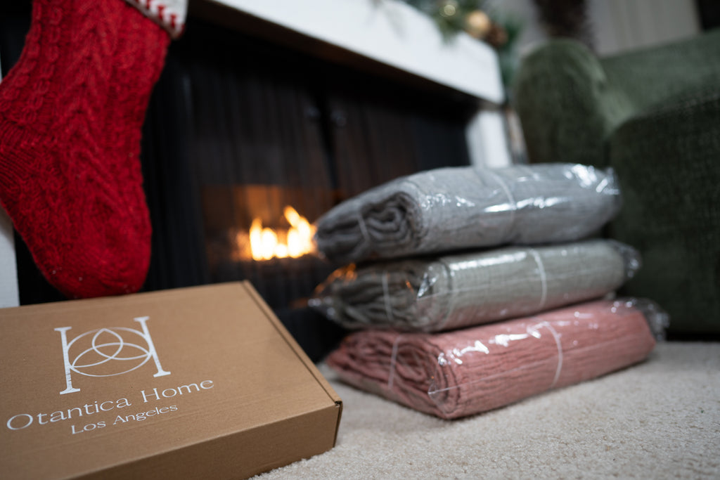 Stack of packaged muslin blankets in blush, sand, and grey colors next to a fireplace and Otantica Home box.