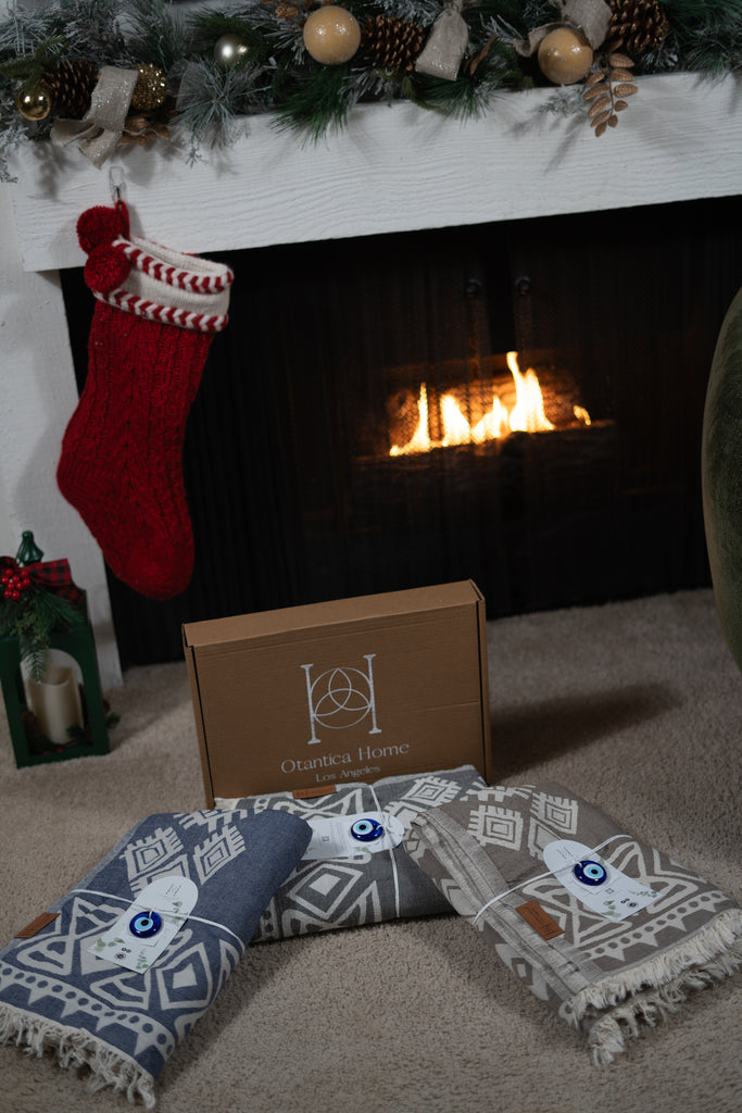 Three folded organic cotton boho blankets placed on a carpet in front of a Christmas fireplace and Otantica Home gift box.