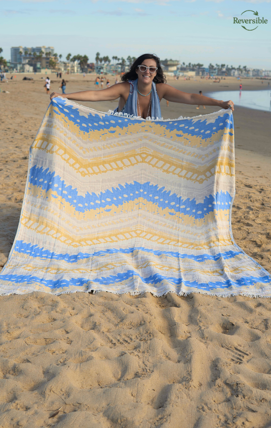 Woman holding a blue and yellow zigzag patterned reversible Havana blanket on a sunny beach.