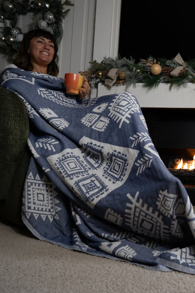 Woman relaxing by the fireplace, wrapped in a dark blue reversible organic cotton boho blanket with geometric patterns.