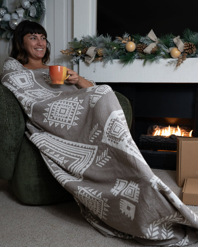 Woman relaxing by the fireplace wrapped in a sand boho organic cotton blanket from Otantica Home