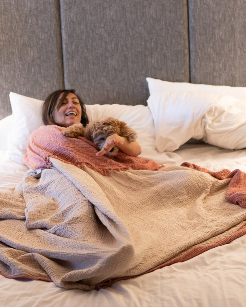 Woman relaxing under blush and sand muslin blankets in a bedroom, displaying reversible double-gauze fabric and comfort.
