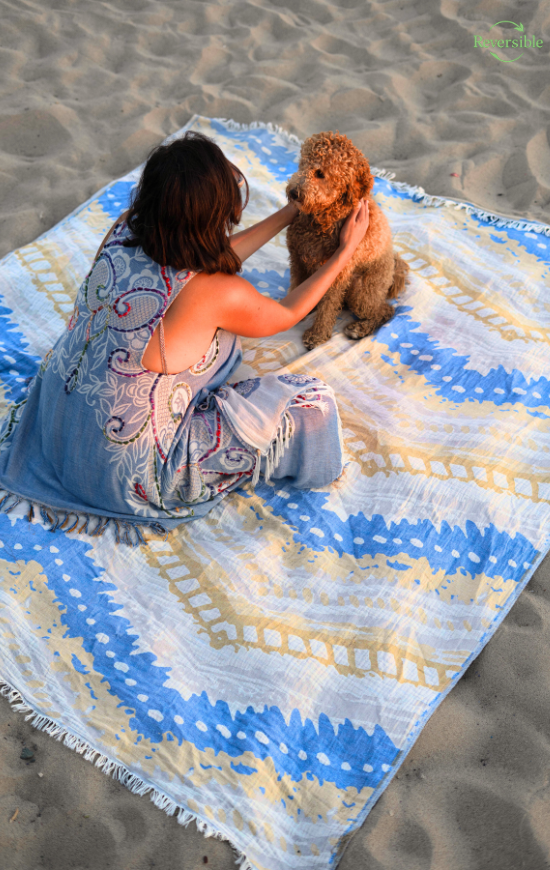 Woman sitting on a sandy beach with her dog on a blue and yellow reversible Havana blanket.