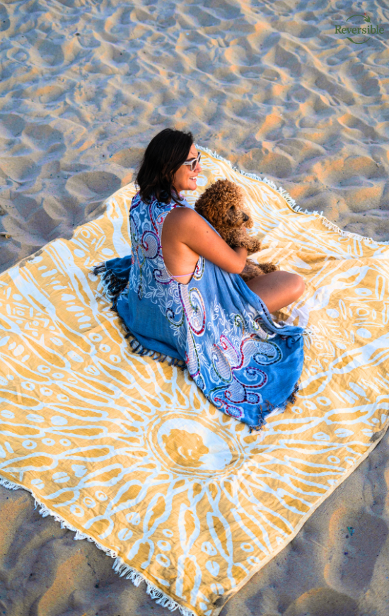 Woman sitting on the sand with her dog on a yellow Sunshine reversible blanket featuring hand-stamped sun patterns on the beach.