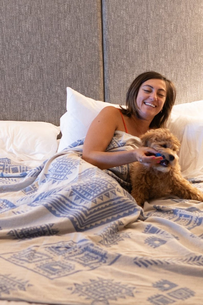 Woman smiling in bed with her dog under a light side of the reversible dark blue boho blanket.
