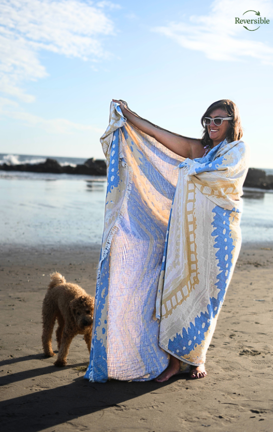 Woman wrapped in a blue and yellow reversible Havana blanket on the beach with a dog walking nearby.