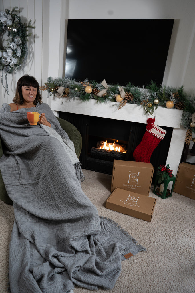 Woman wrapped in a grey muslin blanket drinking from a mug near a fireplace, demonstrating cozy reversible fabric.