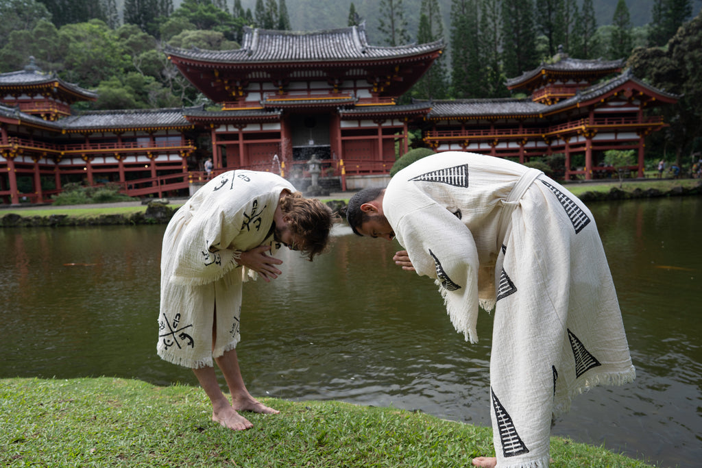 Two men bowing while wearing beige tribal kimonos with black symbols, peaceful ritual-inspired moment